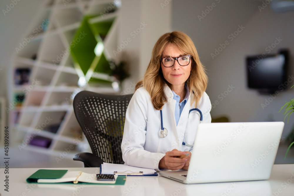 Smiling middle age female doctor sitting at desk and working on laptop at the doctor’s office. Smiling middle age female doctor sitting at desk and working on laptop at the doctor’s office.