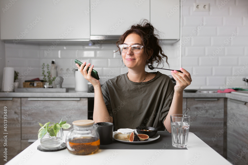 beautiful girl in glasses with curly hair eating breakfast on the kitchen and talking on the phone