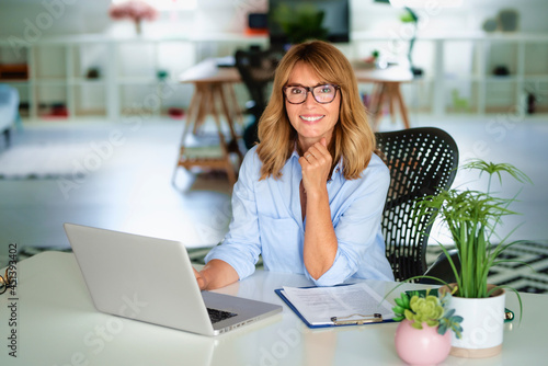 Middle age businesswoman using a laptop while sitting at desk and working at the office. Woman smiling, wearing eyeglasses and shirt