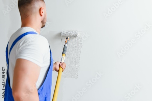 Workman in blue overalls painting the wall with a roller, view from the side