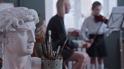 Young woman teacher giving a violin lesson to a little girl - greek man bust and art brushes on the foreground