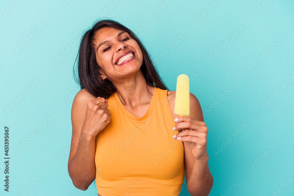 Young latin woman holding ice cream isolated on blue background raising fist after a victory, winner concept. Young latin woman holding ice cream isolated on blue background raising fist after a victory, winner concept.