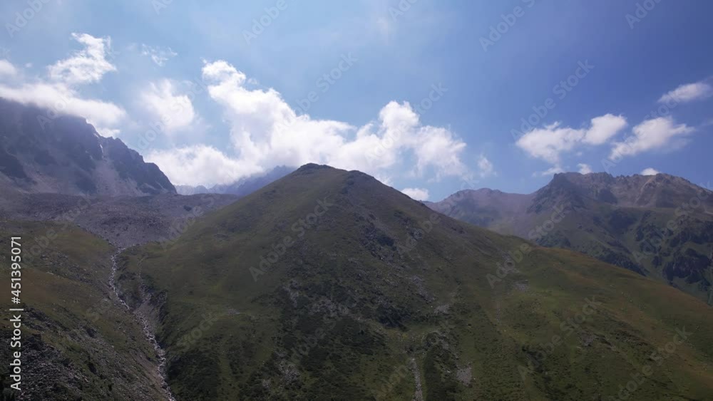 Clouds flow around the tops of high mountains. Rocky gorges and cliffs. The sky is blue, the sun is shining brightly, green grass is growing in places. Mountain terrain from the height of the drone Clouds flow around the tops of high mountains. Rocky gorges and cliffs. The sky is blue, the sun is shining brightly, green grass is growing in places. Mountain terrain from the height of the drone