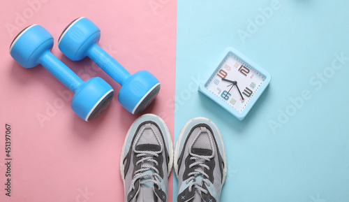 Sports shoes, alarm clock and dumbbells on a blue-pink pastel background. Fitness concept. Top view. Minimalism