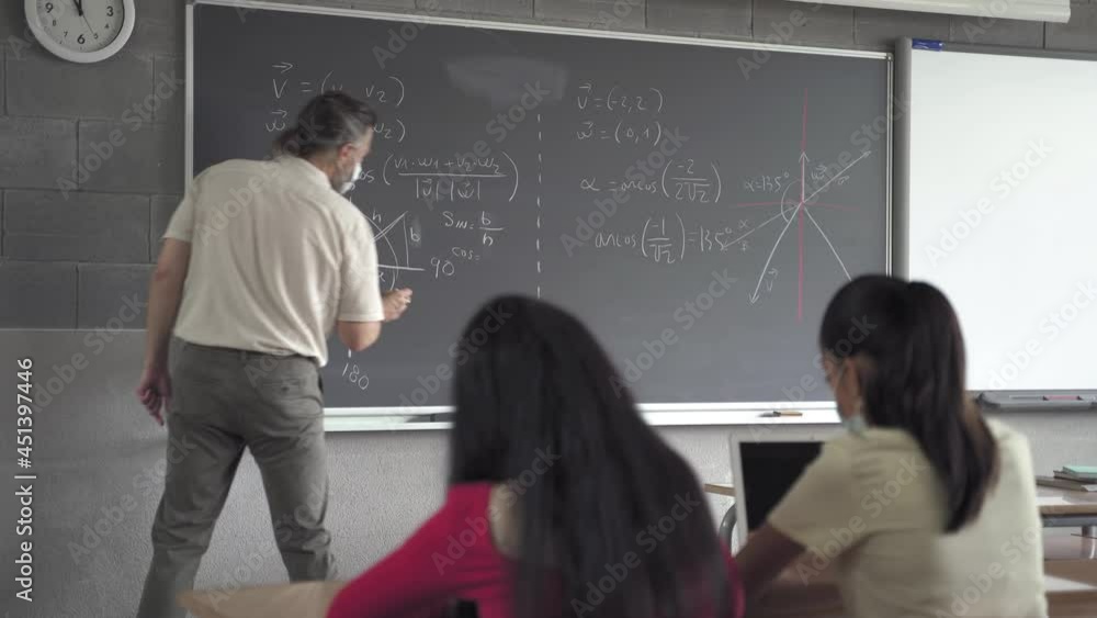 Teacher and students wearing protective face mask in the Secondary School classroom. Professor writing maths formulas and geometry shapes on blackboard Teacher and students wearing protective face mask in the Secondary School classroom. Professor writing maths formulas and geometry shapes on blackboard