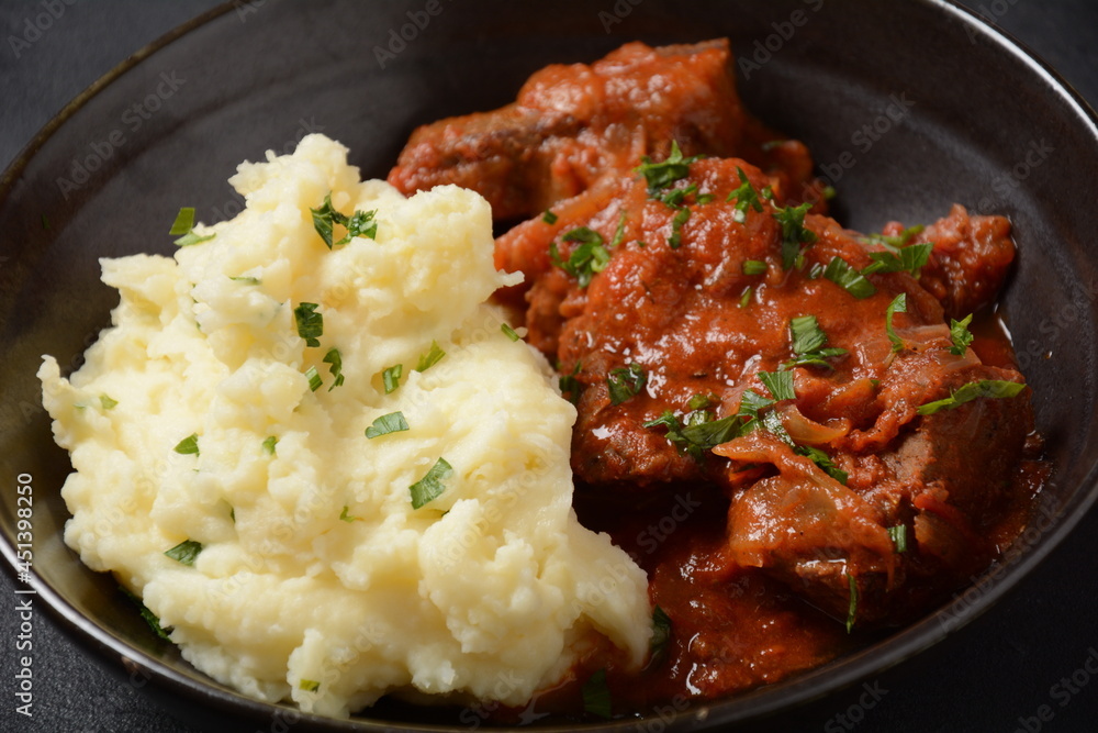 Stewed beef pieces in sauce with mashed potatoes and herbs on dark background Stewed beef pieces in sauce with mashed potatoes and herbs on dark background