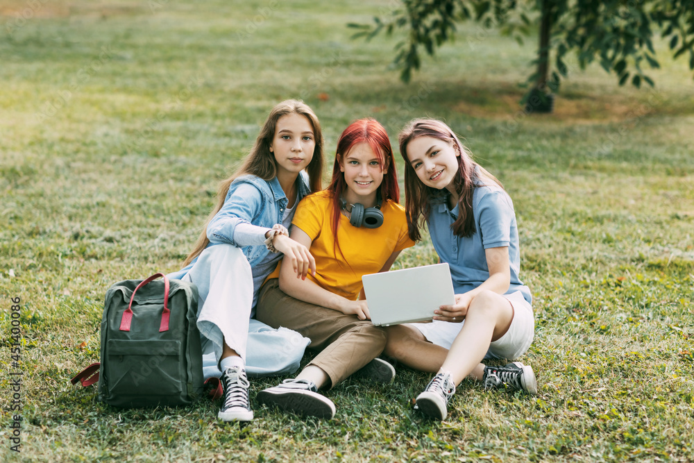 Fototapeta premium Teenage girls are sitting on a green lawn in a park with a backpack and a digital tablet and are preparing a joint project. Education and knowledge, lifestyle of teenagers