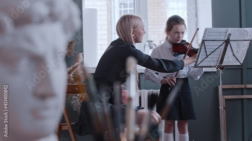 Young woman teacher giving a violin lesson to a little girl in the class - greek man bust on the foreground