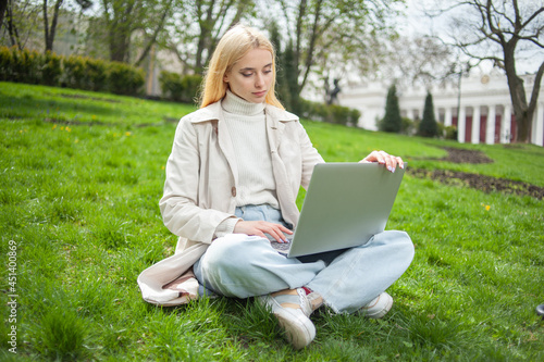 Young blonde woman uses a laptop while sitting on the grass in the park © splitov27