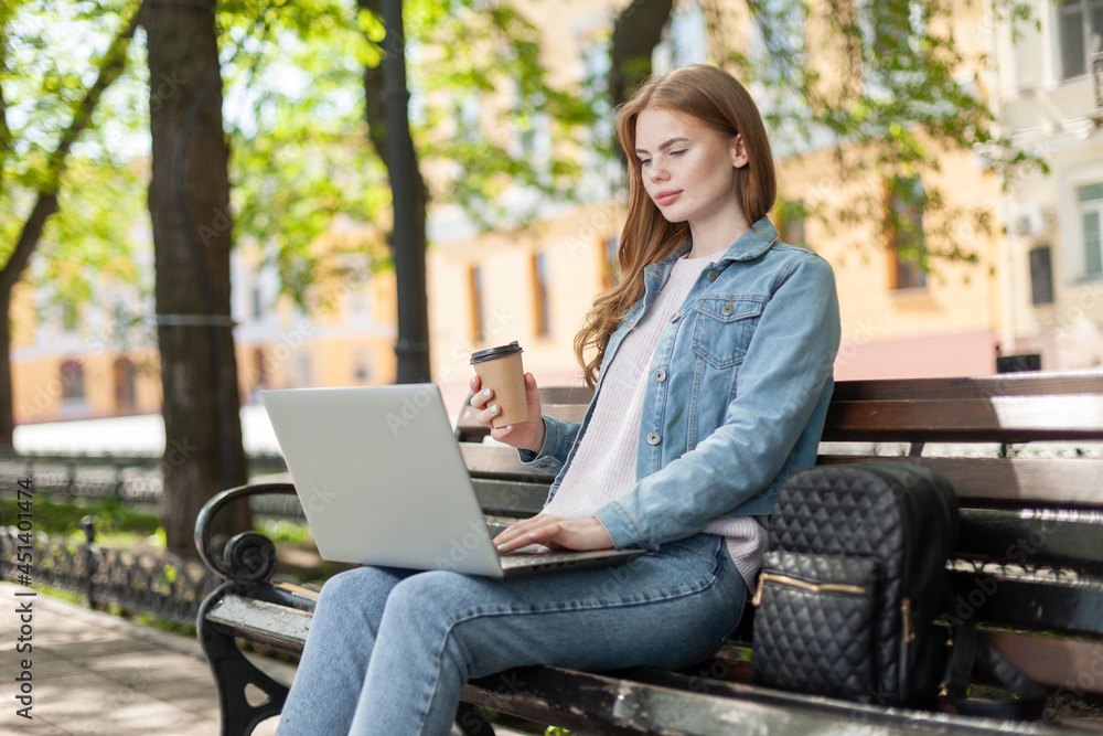 Young pretty woman student or freelancer using laptop while sitting on a bench in the city