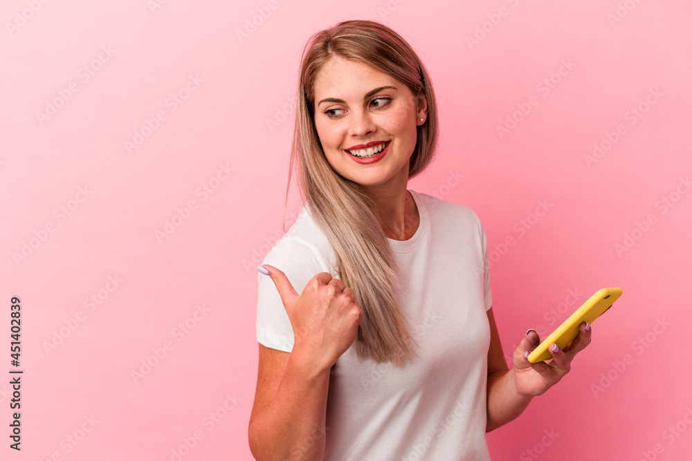 Young russian woman holding a mobile phone isolated on pink background points with thumb finger away, laughing and carefree. Young russian woman holding a mobile phone isolated on pink background points with thumb finger away, laughing and carefree.