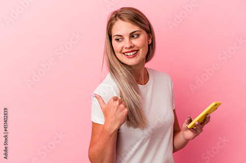 Young russian woman holding a mobile phone isolated on pink background points with thumb finger away, laughing and carefree.