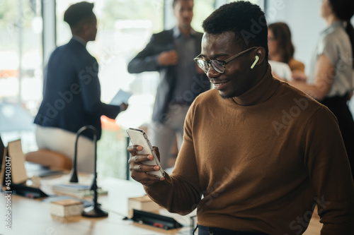 Cheerful African-American Business Man Enjoying Listening to Music on Wireless Earphones and Phone while Leaning on the Table in a Open Plan Office with Multi-Ethnic Team Talking in the Background