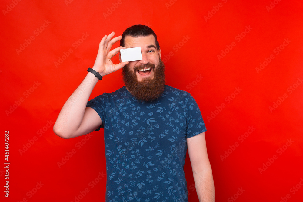 Happy bearded man is covering his eye with a credit or contact card over red background. Happy bearded man is covering his eye with a credit or contact card over red background.