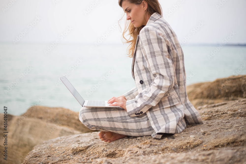 young woman sits on a stone by the sea with a laptop. remote work concept