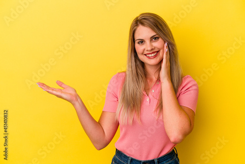 Young russian woman isolated on yellow background holds copy space on a palm, keep hand over cheek. Amazed and delighted.