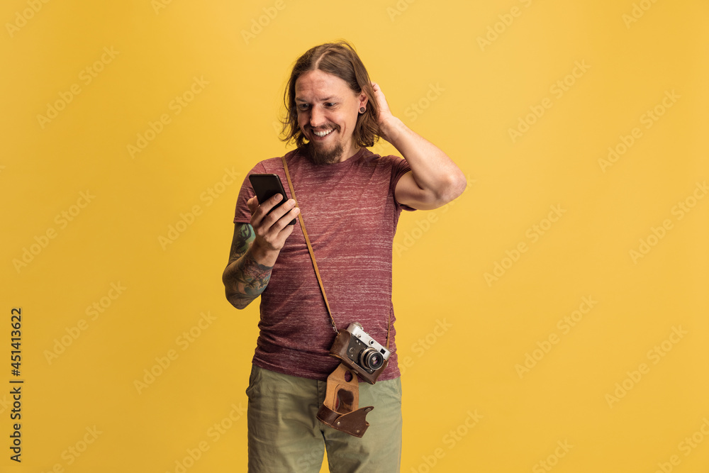 Fototapeta premium Portrait of young man, photographer, cameraman with retro camera using phone isolated on yellow studio background. Concept of occupation, job, funny meme emotions