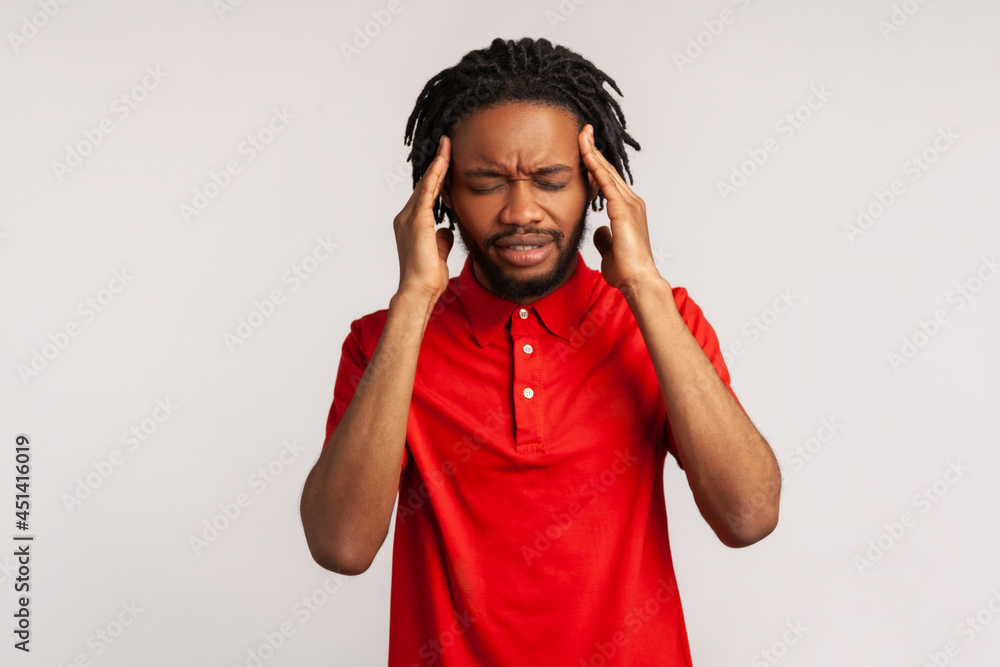 Attractive bearded man with dreadlocks wearing red casual style T-shirt suffering from terrible headache, frowning face, keeping fingers on temples. Indoor studio shot isolated on gray background. Attractive bearded man with dreadlocks wearing red casual style T-shirt suffering from terrible headache, frowning face, keeping fingers on temples. Indoor studio shot isolated on gray background.