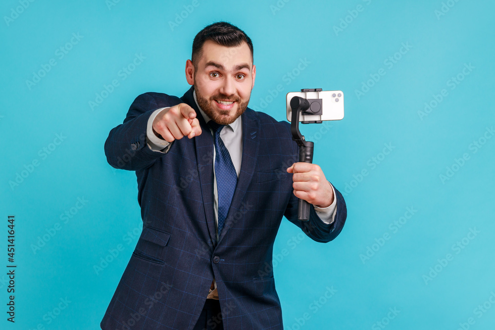 Naklejka premium Smiling blogger man with toothy smile wearing dark official style suit pointing to camera, choosing you, using steadicam for livestreming. Indoor studio shot isolated on blue background.