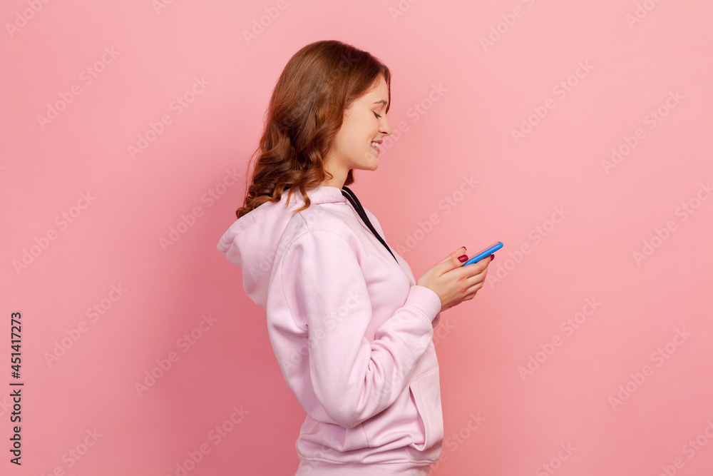 Profile of happy curly haired teenage female in casual hoodie sincerely smiling while using smartphone, messaging, entertainment app. Indoor studio shot isolated on pink background Profile of happy curly haired teenage female in casual hoodie sincerely smiling while using smartphone, messaging, entertainment app. Indoor studio shot isolated on pink background
