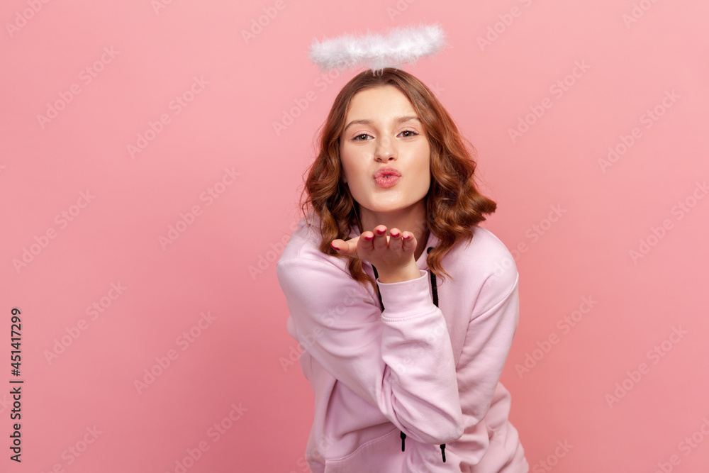 Portrait of joyful curly haired teenage girl in hoodie with halo on head sending air kiss on camera, flirting, valentines day. Indoor studio shot isolated on pink background Portrait of joyful curly haired teenage girl in hoodie with halo on head sending air kiss on camera, flirting, valentines day. Indoor studio shot isolated on pink background