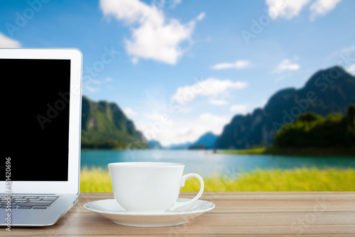 Coffee morning and laptop on wooden table on front of blurred background of the lake, mountain, meadow and blue sky among bright sunlight on a clear day.