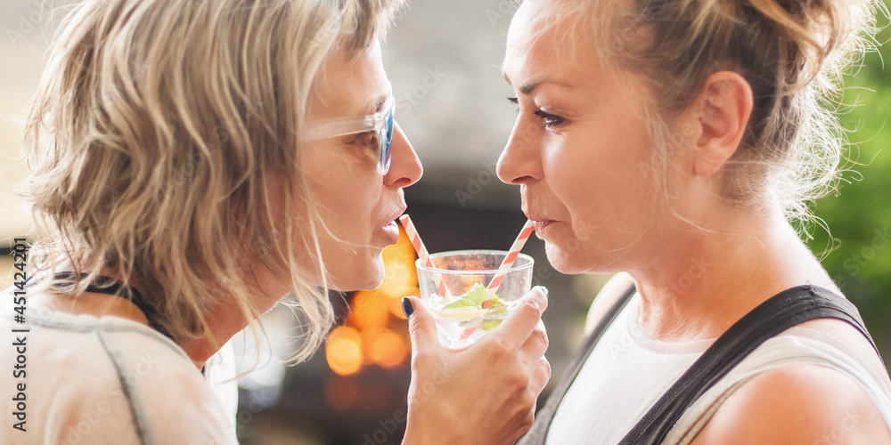 Two cheerful girlfriends drink beverage from one glass using straws, enjoying cold beverage on a summer day outdoors Two cheerful girlfriends drink beverage from one glass using straws, enjoying cold beverage on a summer day outdoors