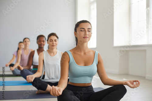 Team of beautiful good looking young women having group meditation class at modern gym or yoga studio, sitting on exercise mats, doing lotus pose, meditating with eyes closed, increasing energy levels