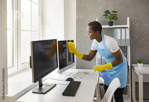 Smiley caretaker, charwoman, daily janitor service lady in uniform with antiseptic spray bottle and wet cloth rag cleaning desktop PC computer, desk and table surfaces in modern office room interior