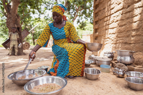 Smiling black African housewife in a beautiful colored dress with matching headscarf, , sitting in her simple outdoor kitchen, preparing a meal photo