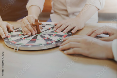 Close up of Young Christian man holding a small wooden cross and hitting in the target center of the dartboard on a wooden table while explains about Jesus story and sharing the gospel to his friends