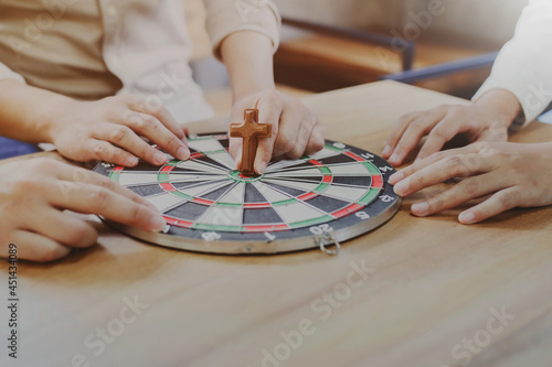 Close up of Young Christian man holding small wooden cross and hitting in the target center of dartboard on wooden table while explains about Jesus story and sharing gospel to his friends 