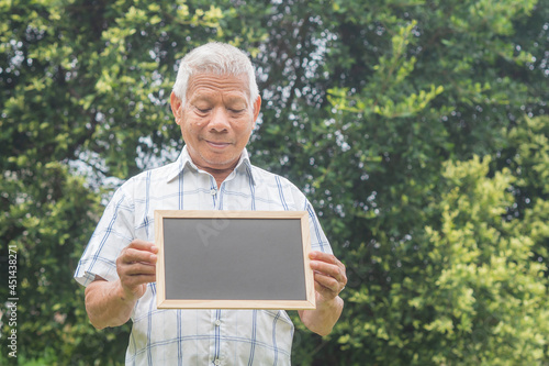 Portrait of a senior man smiling, holding, and looking at a mini blackboard while standing in a garden. Space for text. Aged people and relaxation concept