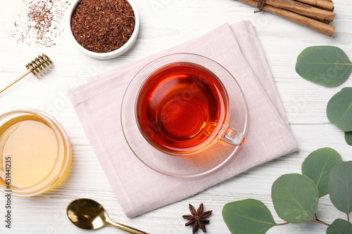 Freshly brewed rooibos tea, dry leaves, honey and spices on white wooden table, flat lay photo