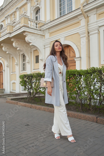 Elegant woman in white trousers and blue oversize blazer. Pretty girl with long wavy brunette hair walking in city