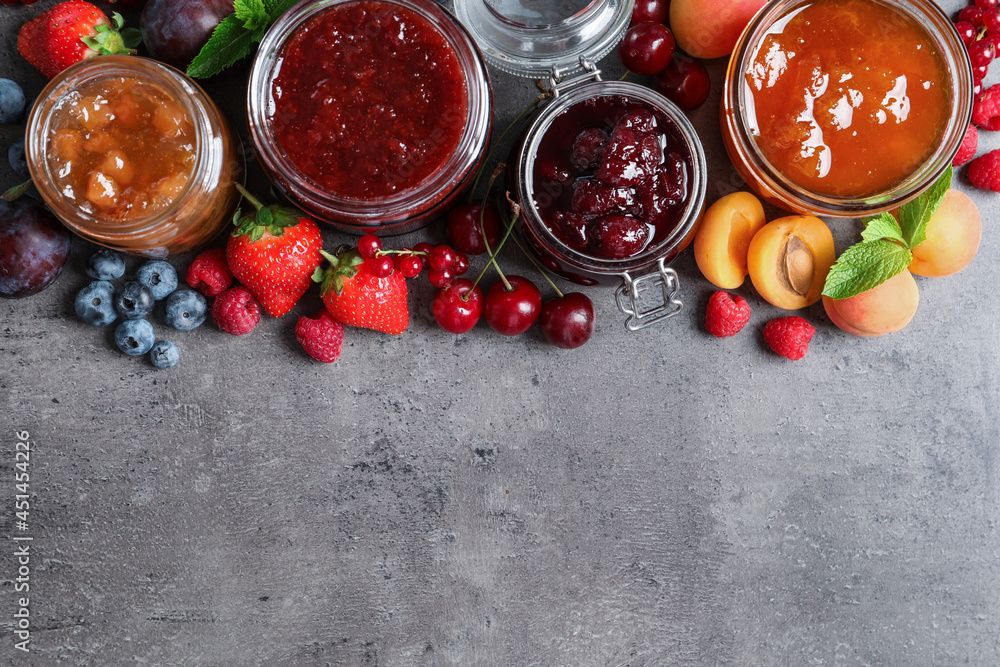 Fototapeta premium Jars with different jams and fresh fruits on grey table, flat lay. Space for text