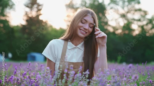 Motion of charming young smiling woman wearing farm outfit sitting in blooming lavender meadow and holding dlower. Front view of girl with long hair enjoying scent of aromatic plant in lavender field. photo