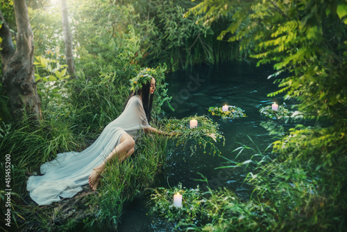 Fabulous mythical natural landscape. Forest fantasy woman sits on shore lake, nymph throws wreath in water. Old Slavic cult ceremony Ivan Kupala. Long white dress. Black hair photo