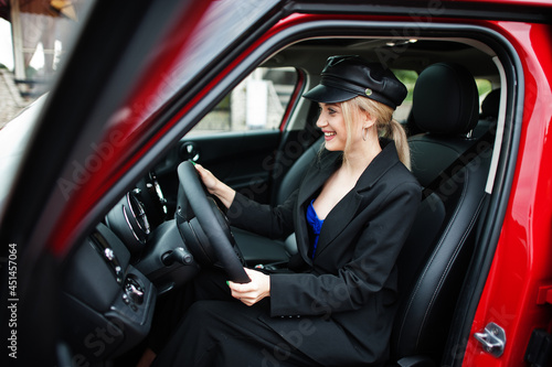 Portrait of beautiful blonde sexy fashion woman model in cap and in all black with bright makeup sit and drive red city car.
