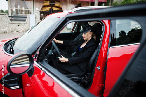 Portrait of beautiful blonde sexy fashion woman model in cap and in all black with bright makeup sit and drive red city car. © AS Photo Family