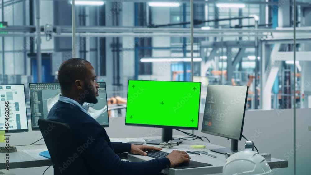 Car Factory Office: Black Engineer Works on a Computer, Two Monitor Screens Show Chroma Key Green Screen and Monitoring of Automated Robot Arm Assembly Line Manufacturing High-Tech Electric Vehicles Car Factory Office: Black Engineer Works on a Computer, Two Monitor Screens Show Chroma Key Green Screen and Monitoring of Automated Robot Arm Assembly Line Manufacturing High-Tech Electric Vehicles