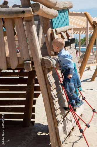 A blond-haired boy climbs a rope ladder on a wooden playground and looks inside. Afternoon walk. Selective focus.