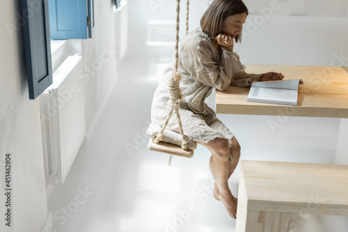 Woman having a leisure time, reading book and sitting on a swing in a bright room. Self-isolation and rest at comfortable home
