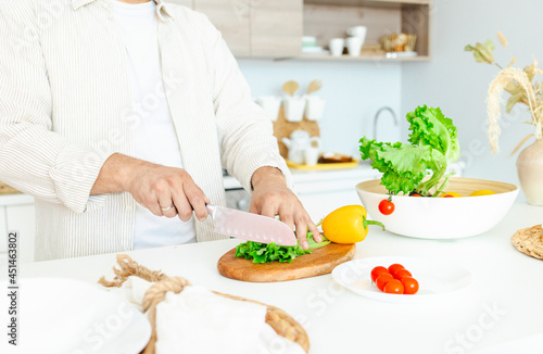 happy young  handsome  bearded man is standing in the modern kitchen prepares a salad of fresh vegetables  cuts vegetables with a knife on a cutting board  cooking as concept of a man s hobby