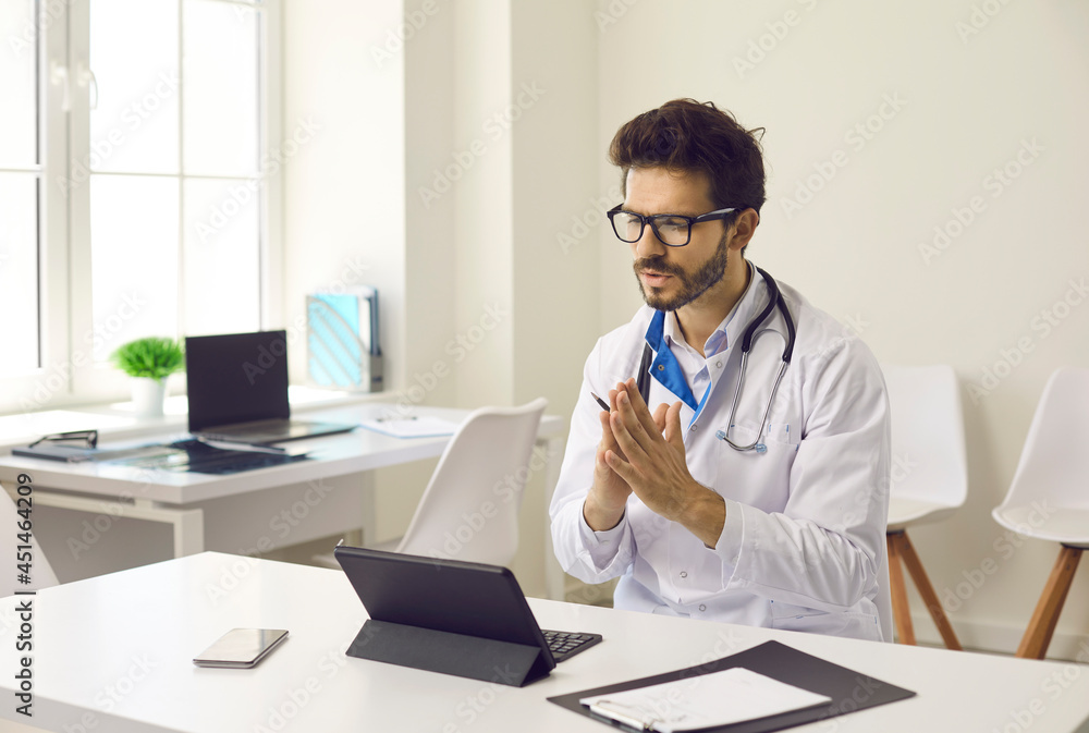 Doctor talking on video call. Serious man in white coat sitting at table with tablet computer device, counseling patient online, explaining how treatment works, discussing and prescribing medication Doctor talking on video call. Serious man in white coat sitting at table with tablet computer device, counseling patient online, explaining how treatment works, discussing and prescribing medication