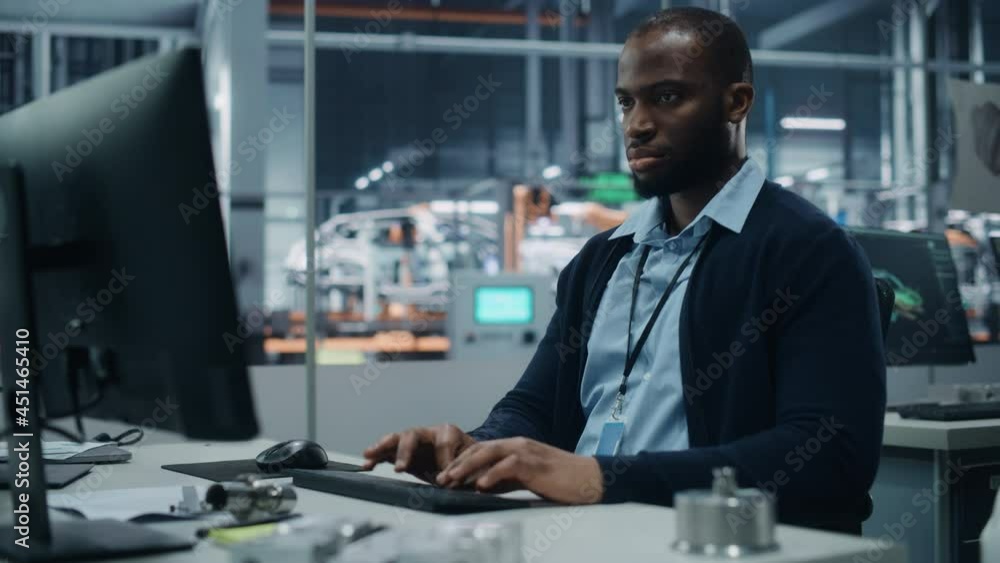 Car Factory Office: Portrait of Confident Black Male Chief Engineer Working on Desktop Computer. Professional Technician in Automated Robot Arm Assembly Line Manufacturing High-Tech Electric Vehicles Car Factory Office: Portrait of Confident Black Male Chief Engineer Working on Desktop Computer. Professional Technician in Automated Robot Arm Assembly Line Manufacturing High-Tech Electric Vehicles