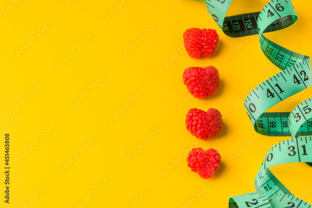 Delicious and juicy raspberries in the form of hearts on a yellow background. Measuring tape for a healthy lifestyle and diet. Summer, freshness and vegetarianism concept. Copy space.