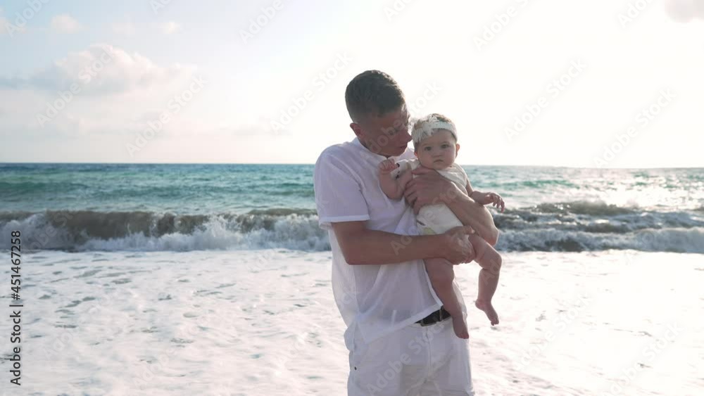 Portrait of young Caucasian father talking to cute little Asian baby daughter with foamy turquoise Mediterranean sea waves rolling at background in sunlight. Man and girl posing at resort at sunset Portrait of young Caucasian father talking to cute little Asian baby daughter with foamy turquoise Mediterranean sea waves rolling at background in sunlight. Man and girl posing at resort at sunset