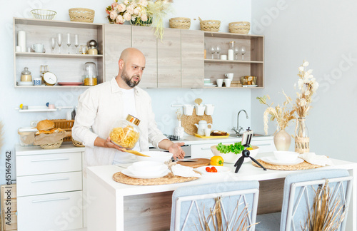happy young, handsome, bearded man is standing in the modern kitchen with pasta in his hands and vegetables on table, cooking as concept of a man's hobby, records video for food blog