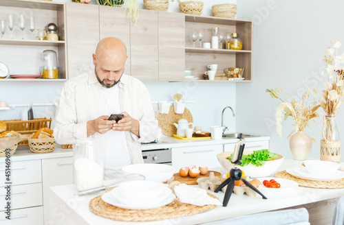 happy young, handsome, bearded man is standing in modern kitchen and talking on phone before prepares a salad of fresh vegetables lettuce, tomatoes, sweet pepper and eggs, concept of culinary blog